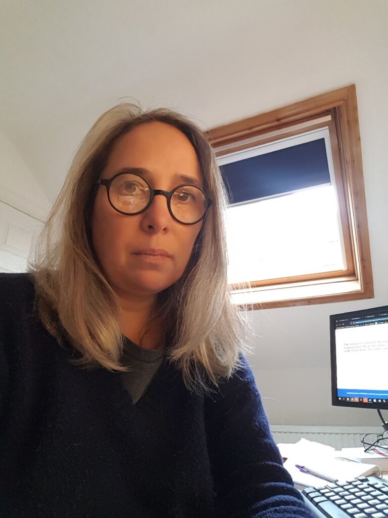 Julia Molinari looking bemused into the camera sitting at her office desk with a window and computer screen behind her.